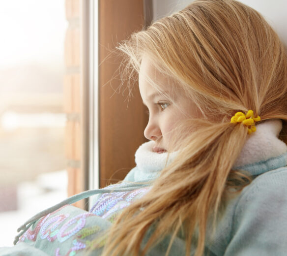 Sad female child with blonde hair sitting on windowsill, looking out window with unhappy expressionfeeling lonely
